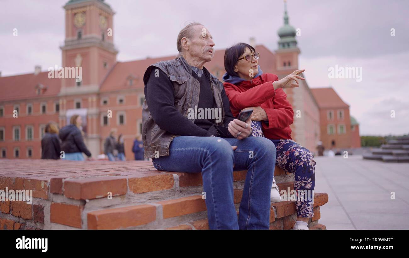 Ältere Touristen sitzen und unterhalten sich im historischen Zentrum einer alten europäischen Stadt. Senior hat ein Smartphone in der Hand. Palast Stockfoto