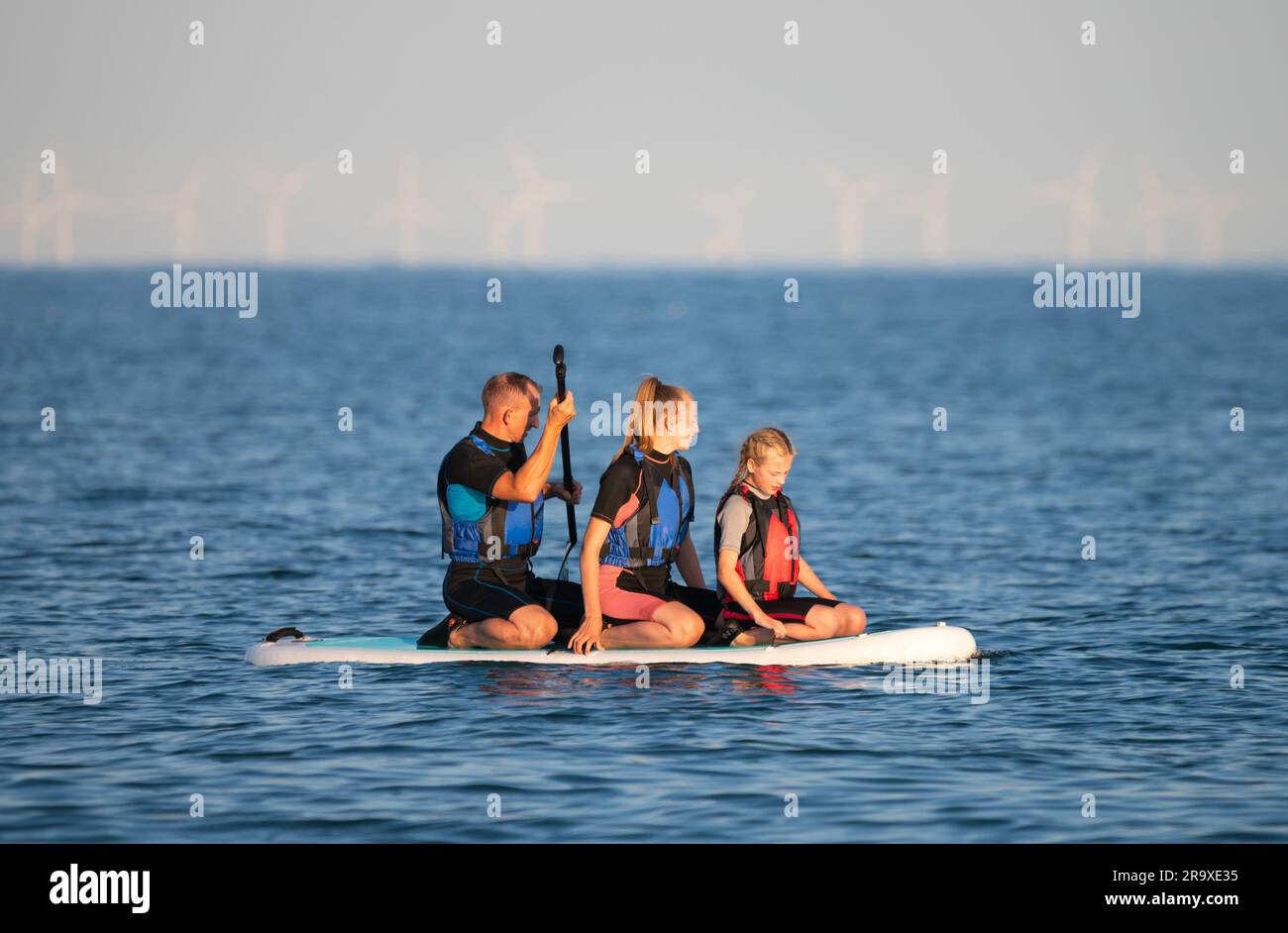 Männlicher Erwachsener mit 2 jüngeren Frauen (möglicherweise eine Familie) auf einem Paddleboard auf dem Meer, sitzt während des Paddelns an einem heißen Sommertag in England, Großbritannien. Stockfoto