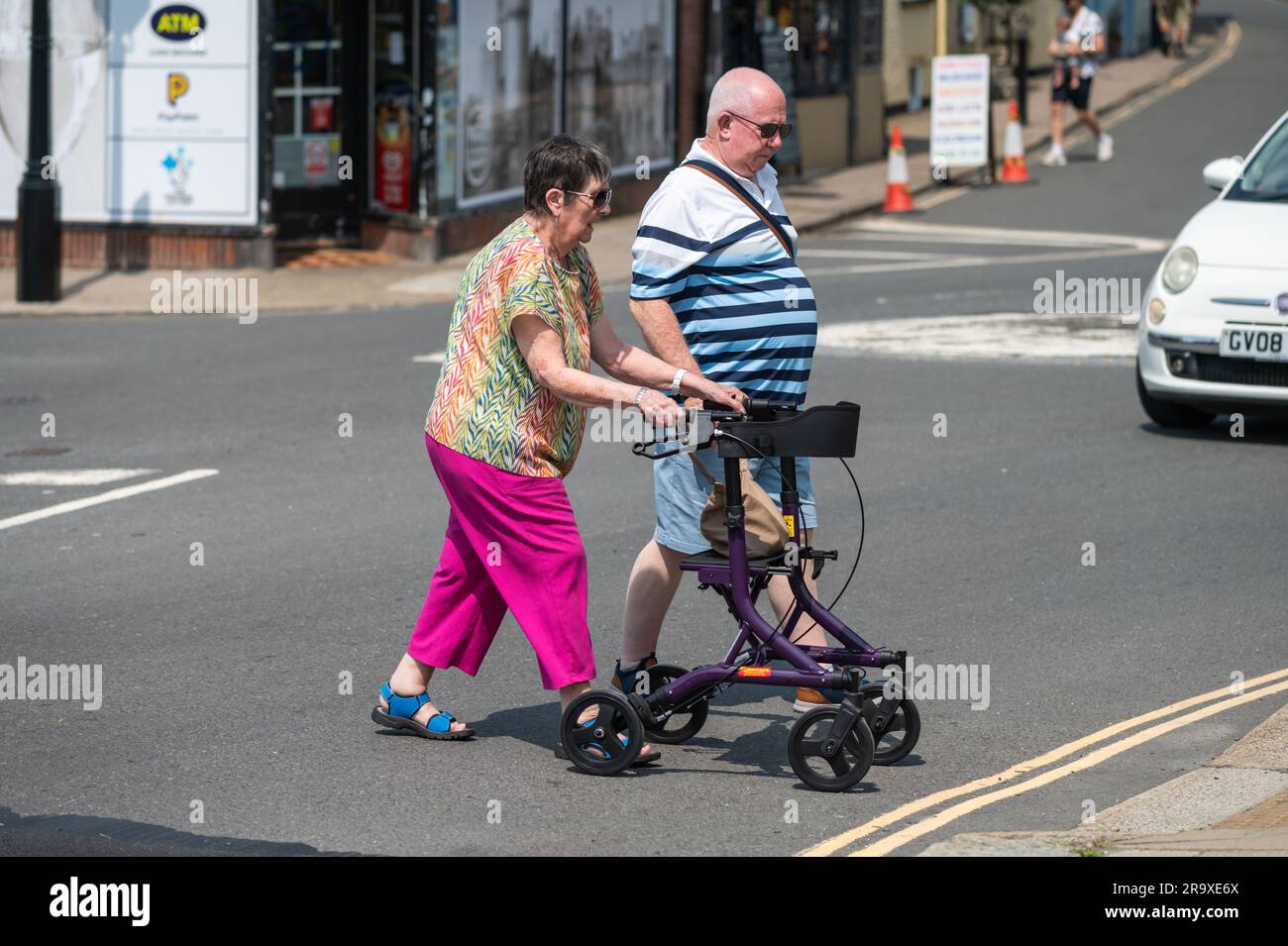 Frau, die mit Hilfe eines Rollers oder Rollers auf Rädern eine Straße überquert, eine Gehhilfe für behinderte Menschen in England, Großbritannien. Stockfoto