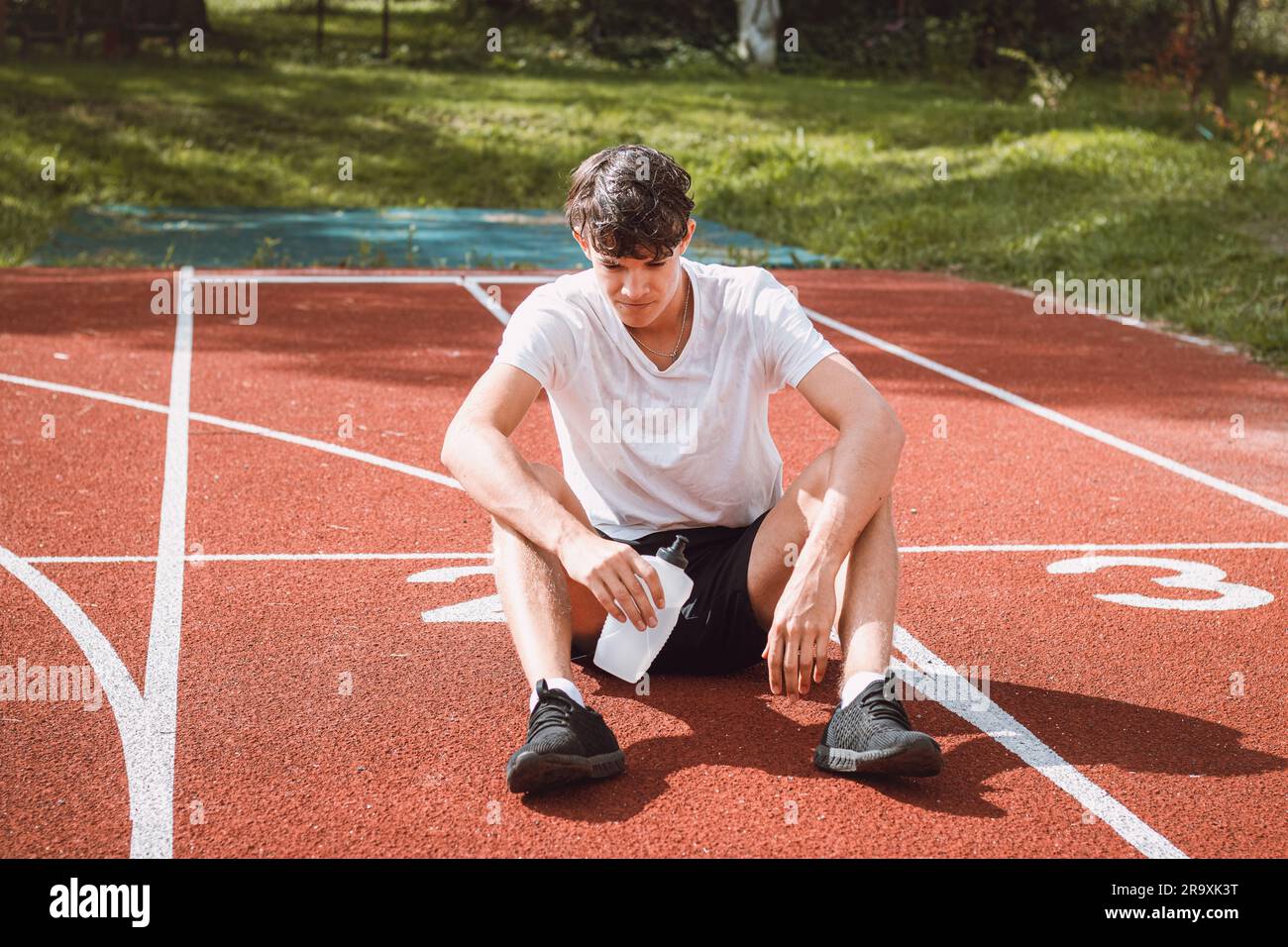 Der junge Sportler erfrischt sich nach einem harten Training auf dem sportlichen Oval in der intensiven Hitze mit Wasser. Ausdauertraining. Braunhaariger Jugendlicher. Stockfoto