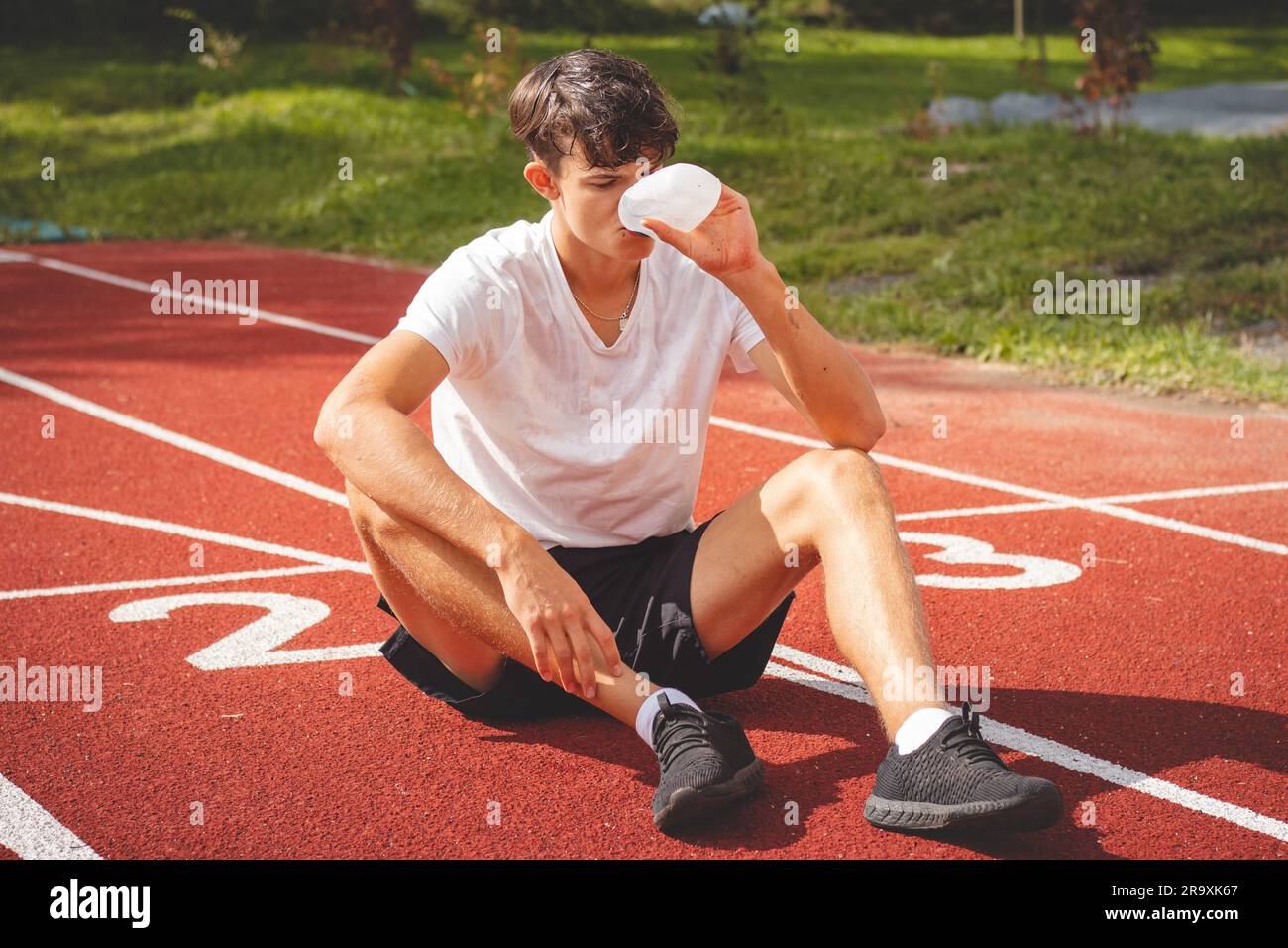Der junge Sportler erfrischt sich nach einem harten Training auf dem sportlichen Oval in der intensiven Hitze mit Wasser. Ausdauertraining. Braunhaariger Jugendlicher. Stockfoto