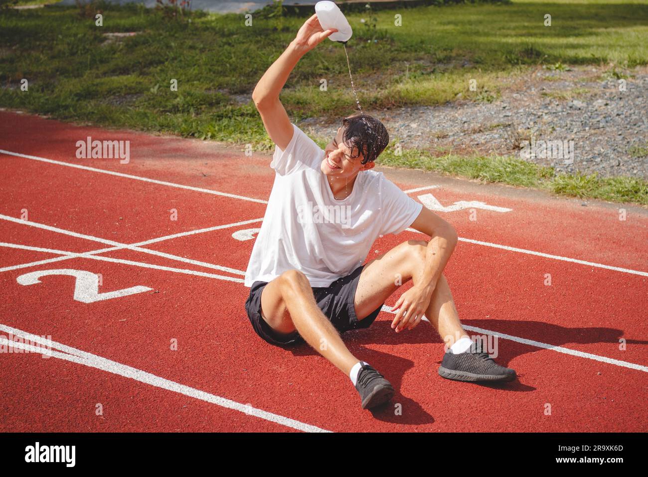 Der junge Sportler erfrischt sich nach einem harten Training auf dem sportlichen Oval in der intensiven Hitze mit Wasser. Ausdauertraining. Braunhaariger Jugendlicher. Stockfoto