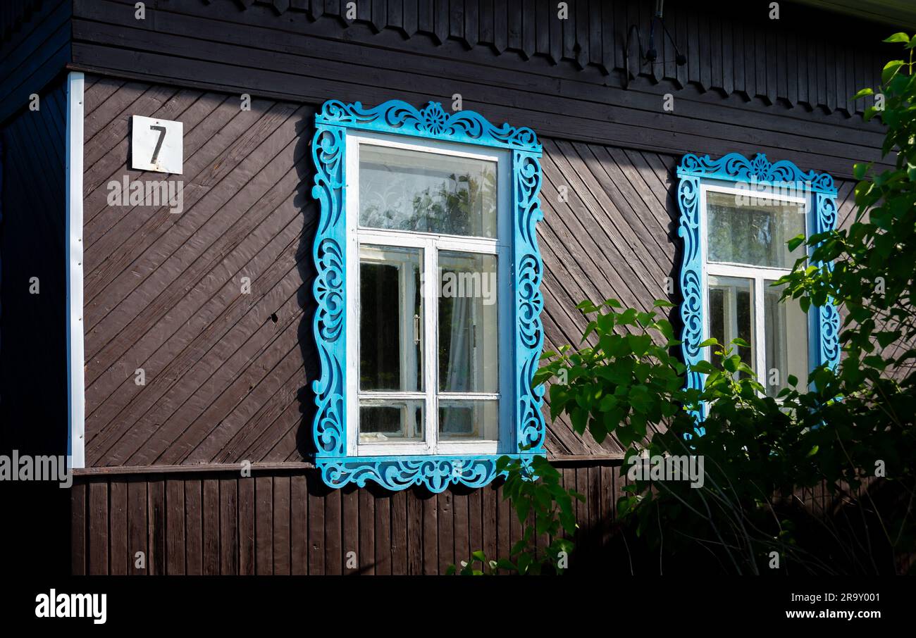 Zwei Fenster mit blau geschnitzten Holzjammen, die mit traditionellen Mustern an der Fassade eines russischen isba, einem aus Holz erbauten Haus in Russland, dekoriert sind Stockfoto