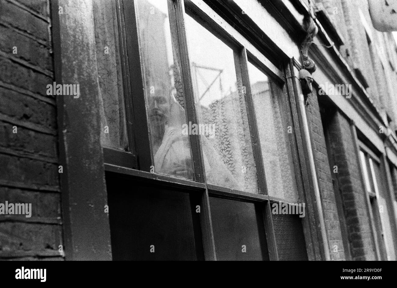 Whitechapel London in einer Straße gleich neben der Brick Lane starrt eine einsame asiatische Frau aus ihrem flachen Fenster. Es gab Berichte in der Presse, dass viele neue Einwanderer nach Großbritannien, insbesondere Frauen aus Bangladesch, von ihren Männern unter Verschluss gehalten wurden. Sie sprachen die Sprache nicht und durften selten ihre Häuser verlassen. Whitechapel, London, England 1978. Stockfoto
