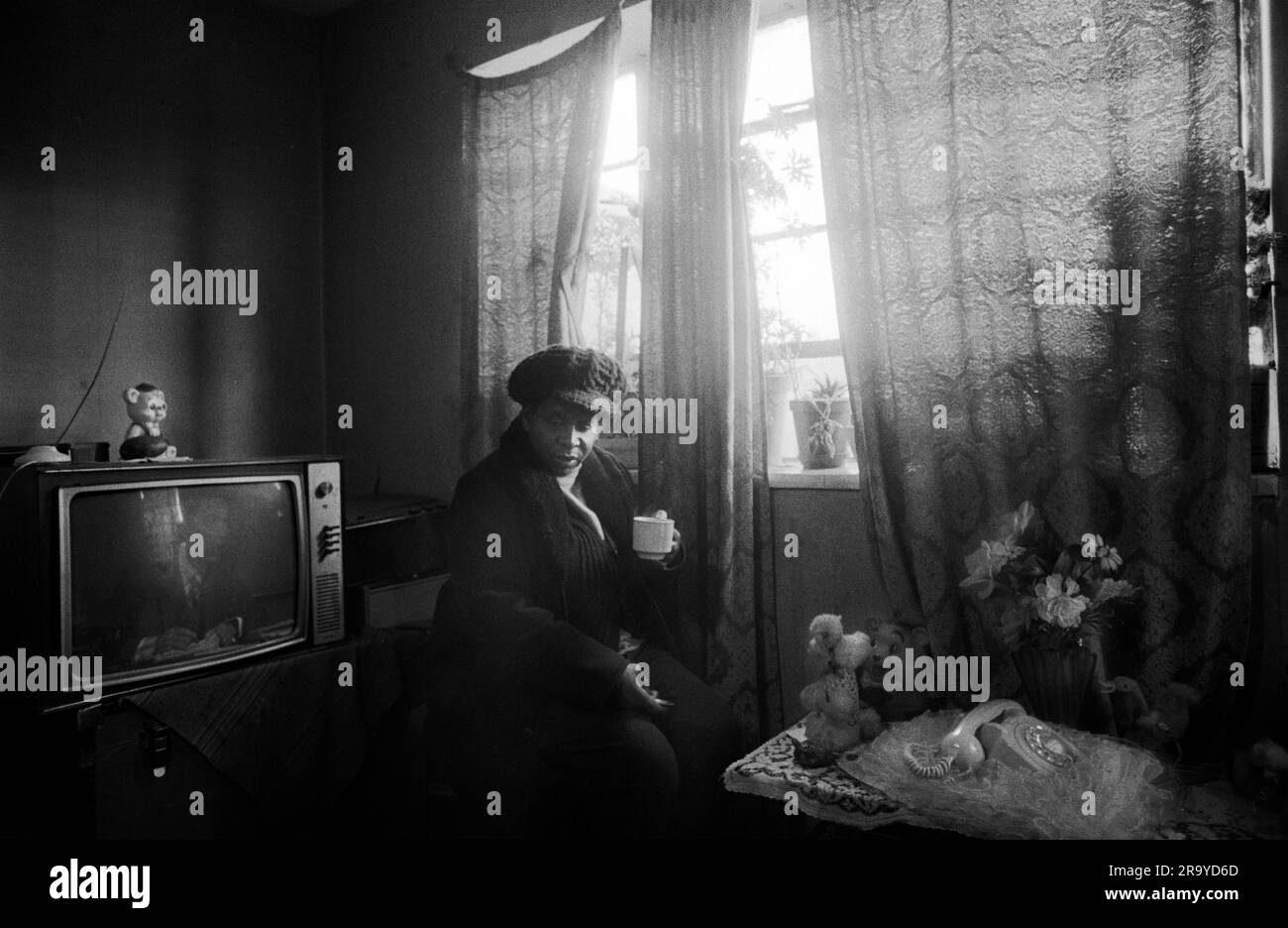 East End London 1970er, Armut in Großbritannien, eine ältere schwarze britische Frau in ihrem wohnsitz auf dem Stonebridge Estate. Isoliert und allein in einem Einzelzimmer plus kleine Küche und Bad. Alles in sehr schlechtem Zustand, die Wohnung ist feucht und sehr kalt, sie trägt Hut und Mantel und trinkt eine Tasse Tee, um sich warm zu halten. Haggerston, East London, England 1978. 70S GB HOMER SYKES Stockfoto