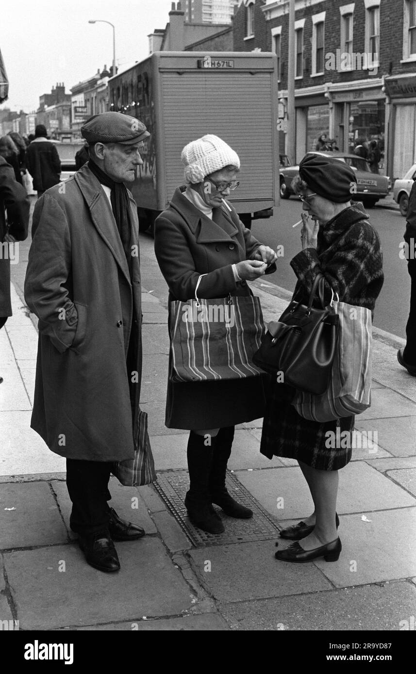 East End London 1970er Roman Road Market. Zwei Arbeiterinnen auf der Straße leuchten auf. Eine schlägt ein Streichholz aus einer Schachtel Streichhölzer für die anderen Zigaretten, während ihr Mann zusieht. Er trägt die Einkaufstasche und trägt eine flache Mütze und einen Schal, die auf traditionelle Weise getragen und gefesselt werden. Sie gehen einkaufen auf dem Roman Road Markt. Tower Hamlets, London, England um 1975. 70S GB HOMER SYKES Stockfoto