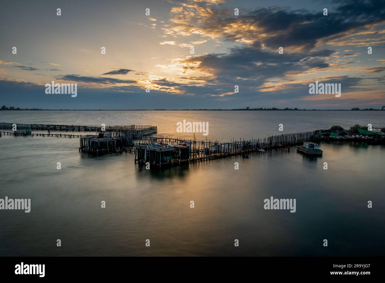 Lange Exposition einer Fischfarm bei Sonnenuntergang mit der Sonne, die die Wolken erhellt und Licht auf das Wasser wirft. Stockfoto