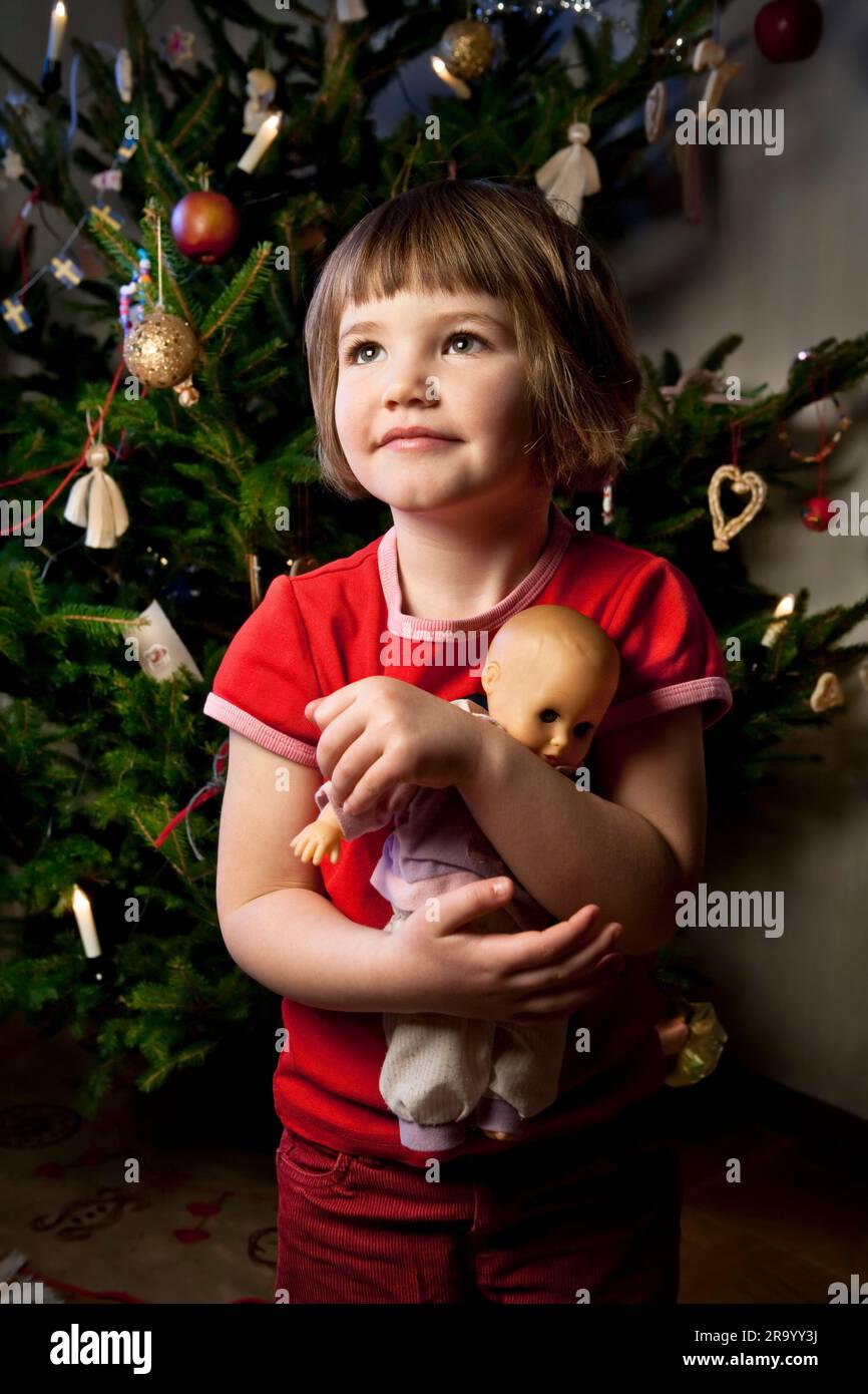 Kleines Mädchen mit seiner Puppe wegschauen vor einem Weihnachtsbaum Stockfoto