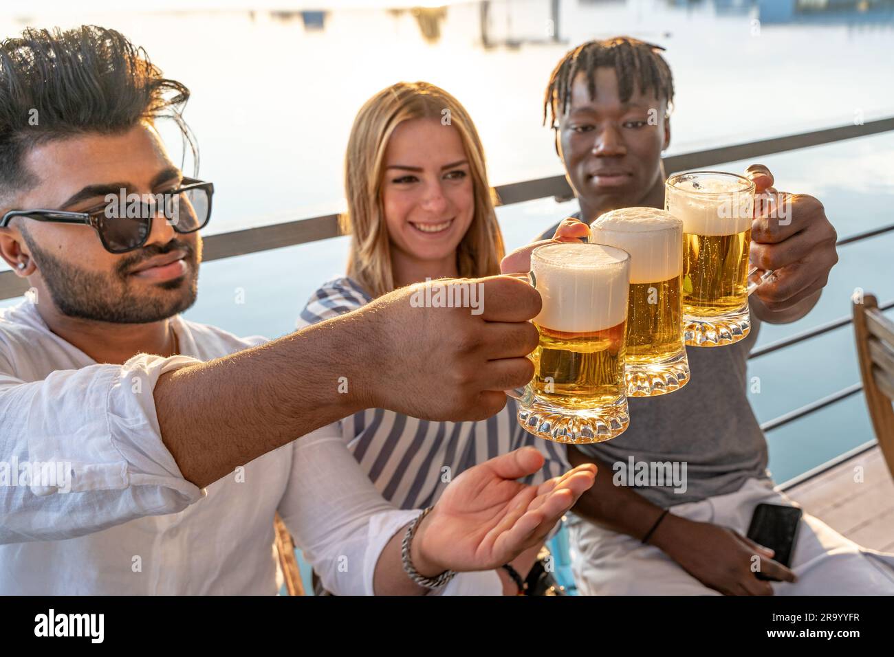 Kleine Gruppe von jungen erwachsenen multiethnischen Freunden, die mit drei Tassen Bier anstoßen - Sommerferienkonzept auf einem Schiff auf dem Fluss Stockfoto