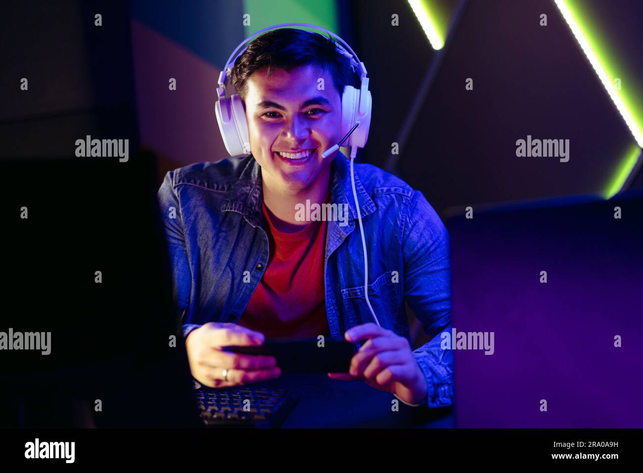 Ein Gamer, der mit einem Headset und einem Smartphone in der Hand die Kamera anlächelt. Ein junger männlicher Spieler sitzt an seiner Spielstation und sieht gern live zu Stockfoto
