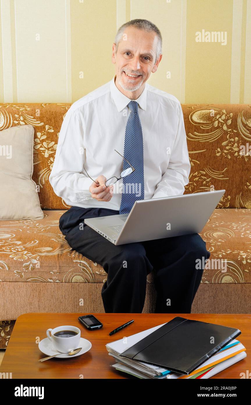 Ein gutaussehender Geschäftsmann, der zu Hause mit einem Computer arbeitet, eine Brille in der Hand hält und bereit ist, eine heiße Tasse schwarzen Kaffee zu trinken Stockfoto