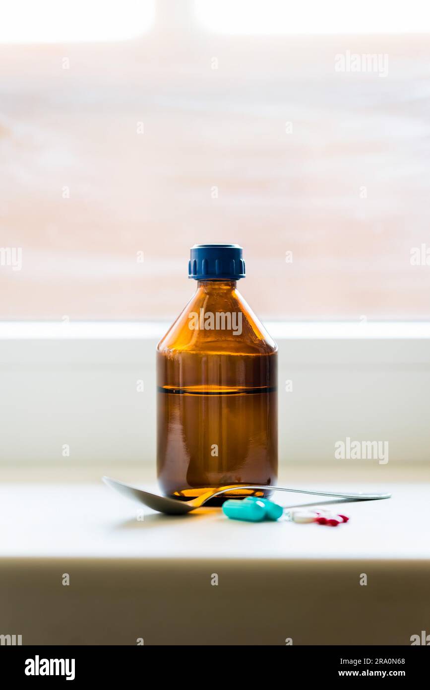 Eine Flasche Hustensaft mit einem Löffel und einige Pillen in der Nähe des Fensters Stockfoto