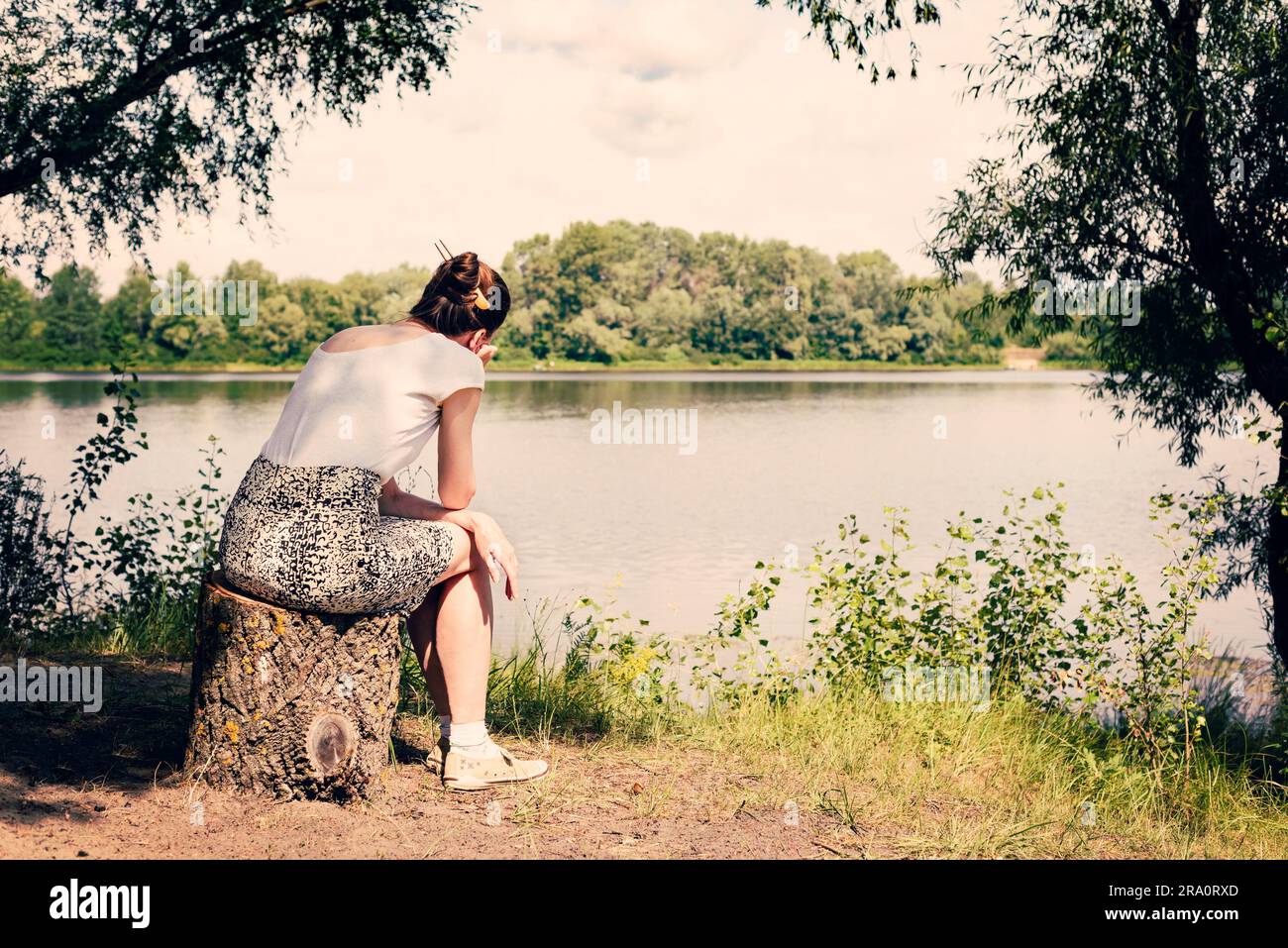 Eine traurige und nostalgische Frau sitzt in der Nähe des Dnieper-Flusses in Kiew, Ukraine. Sie denkt, während sie zusieht oder weit in der Ferne beobachtet Stockfoto