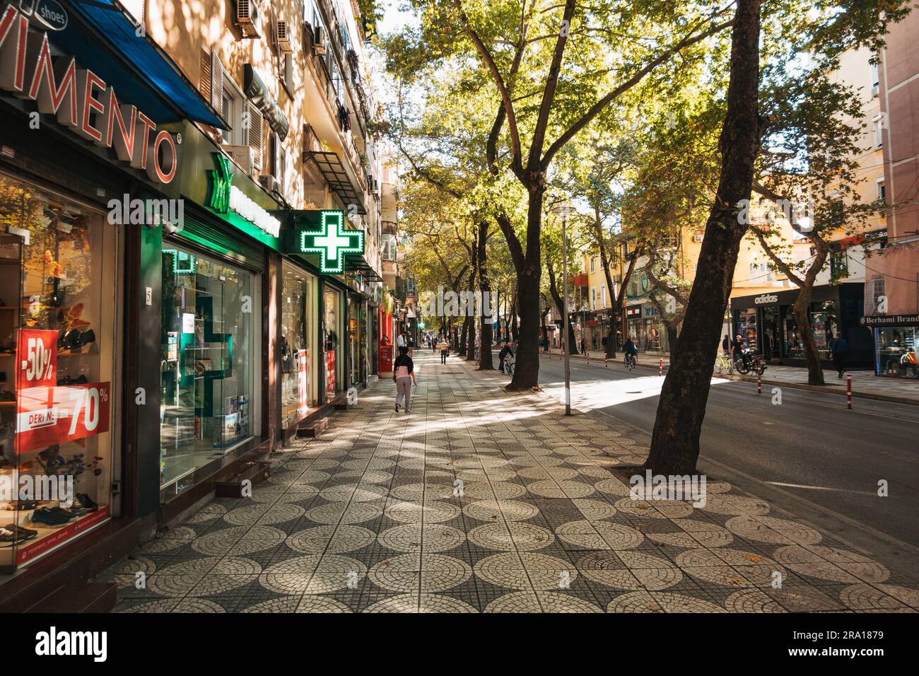 Geschäfte entlang einer schattigen Stadtstraße in Tirana, Albanien, mit gemusterten Fliesen auf dem Bürgersteig und Bäumen, die einer westeuropäischen Stadt ähneln Stockfoto
