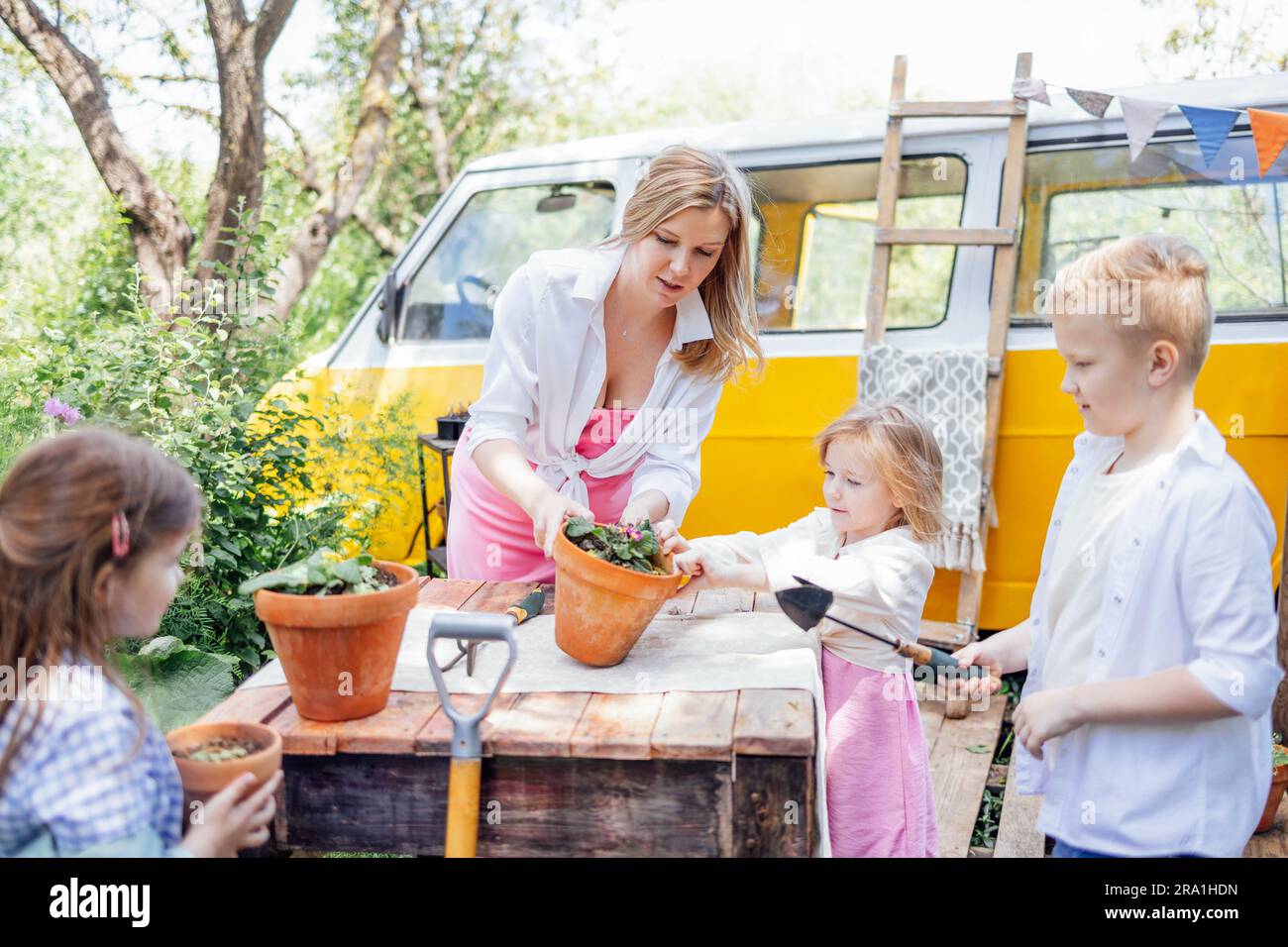 Eine junge Frau und ihre drei Kinder Pflanzen Blumen in Töpfen im Garten. Hübsche Mutter, süße Mädchen und Junge sind im Gartenbau beschäftigt. Glückliche Familie Stockfoto