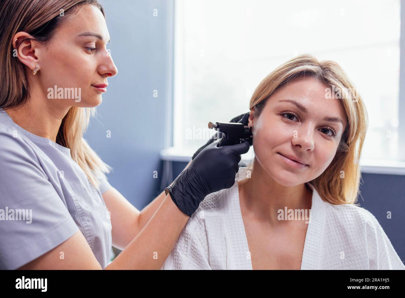 Nahaufnahme des Gesichts einer jungen blonden Frau, die eine Ohrpiercing-Operation durchführt. Arzthandschuhe in schwarzen Handschuhen halten eine Ohrstechpistole. Medizinisches Personal macht Loch an Stockfoto