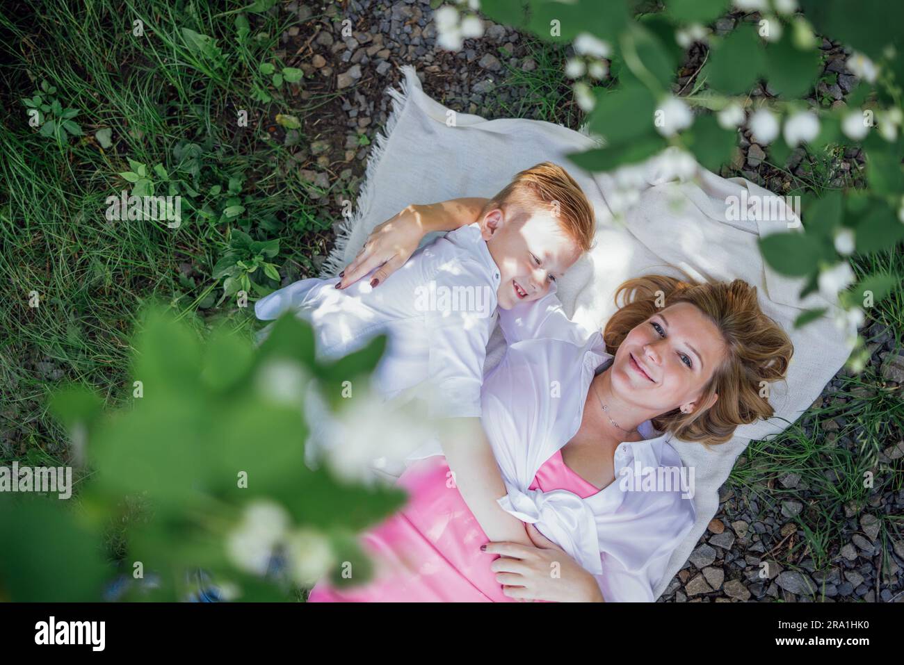 Die junge Mutter und ihr Sohn umarmen sich und liegen auf einer Decke auf dem Gras. Eine attraktive Frau und ein lustiger Junge lächeln und haben im Garten Spaß. Anzeigen Stockfoto