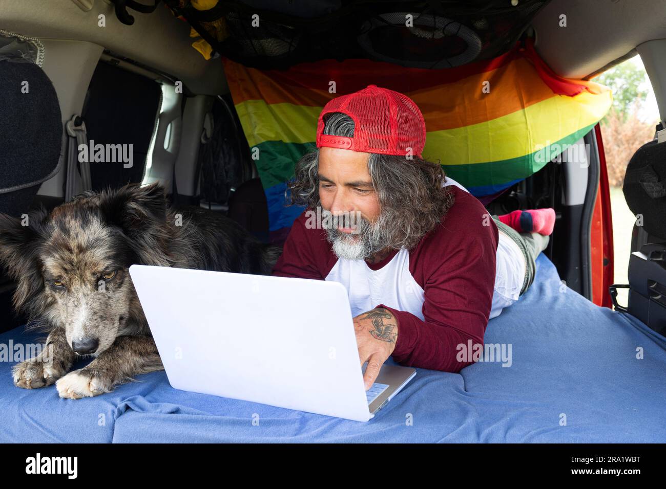 Ein Mann mittleren Alters, der mit seinem Haustier im Campingwagen liegt und einen Laptop benutzt. Ein Reisender mit einem Remote-Computer, der online arbeitet und Spaß hat Stockfoto