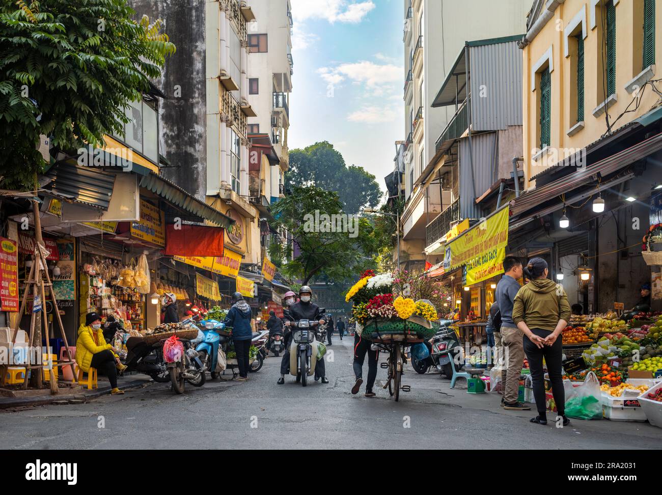 Eine Blumenverkäuferin schubst ihr beladenes Fahrrad vorbei an geschäftigen Geschäften in Cau Go, in der Altstadt von Hanoi, Vietnam. Stockfoto