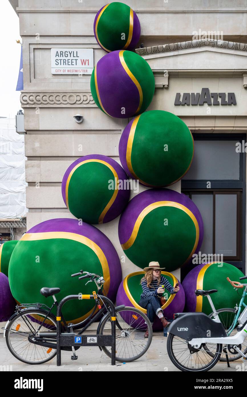 London, Großbritannien. 30. Juni 2023 Eine Frau sitzt mit einem Kaffee neben riesigen, von Wimbledon inspirierten Tennisballdekorationen vor dem Lavazza-Laden in der Argyll Street. Das jährliche zweiwöchige Tennisturnier in Wimbledon beginnt nächste Woche. Kredit: Stephen Chung / Alamy Live News Stockfoto
