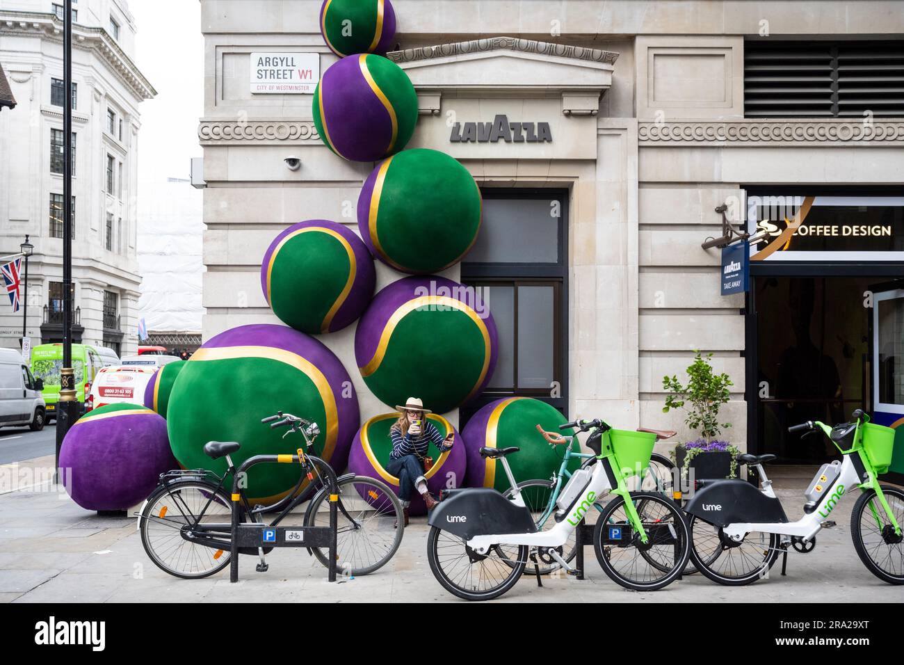London, Großbritannien. 30. Juni 2023 Eine Frau sitzt mit einem Kaffee neben riesigen, von Wimbledon inspirierten Tennisballdekorationen vor dem Lavazza-Laden in der Argyll Street. Das jährliche zweiwöchige Tennisturnier in Wimbledon beginnt nächste Woche. Kredit: Stephen Chung / Alamy Live News Stockfoto