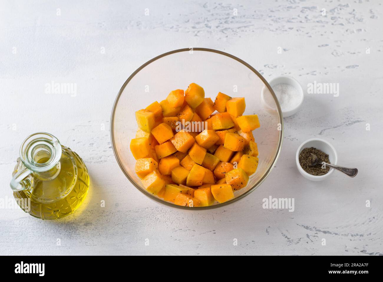 Glasschale mit gewürfeltem Kürbis, Olivenöl, Salz und Pfeffer auf einem hellblauen Tisch, Draufsicht. Köstlicher veganischer Auflauf. Stockfoto