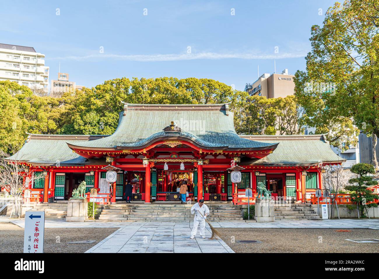 Die Halle, Haiden, des Shinto-Ikuta-Schreins in Kobe, Japan. Zinnrot und grüner Saal mit Bäumen und blauem Himmel dahinter, ein Mann fegt Blätter vor sich. Stockfoto