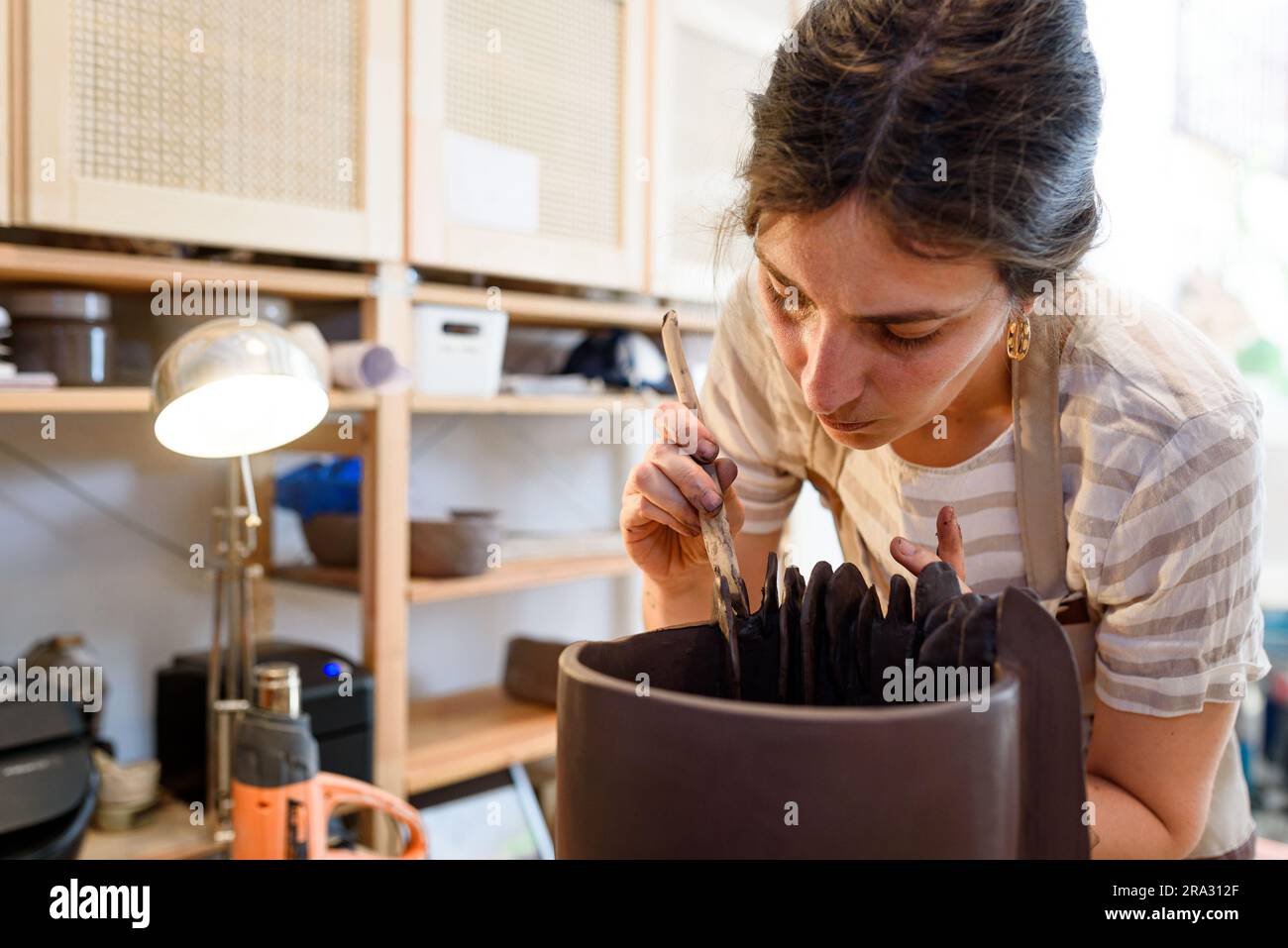 Eine Frau in einer Töpferwerkstatt, die rohe Keramikvase und Lehm bearbeitet, um Gras zu machen. Keramikwerkstatt mit natürlichem Licht zum Schaben, Glätten, Formen und Formen Stockfoto