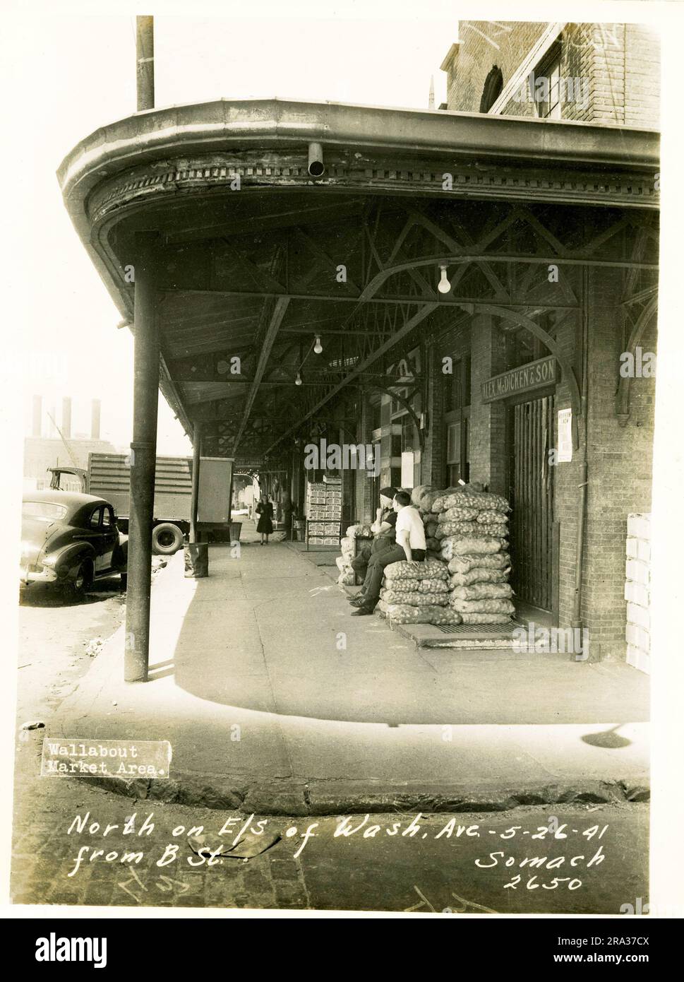 Foto von der Fassade des Wallabout Market, nördlich auf der E/S der Washington Ave. Von der B Street. Blick auf die Straße von der Washington Avenue Ecke mit Blick auf den Bürgersteig entlang des McDicken Ladengeschäfts, wo Männer auf Säcken sitzen und eine Frau auf sie zuschauen, von Negativ 2650... Stockfoto