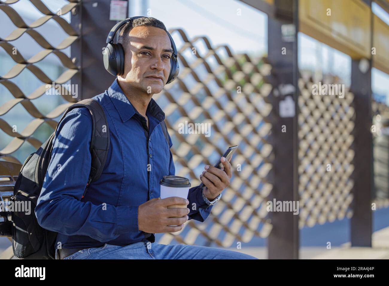 Junger lateiner mit Kopfhörern, der auf den Bus wartet, mit einer Papptasse Kaffee in der Hand und Kopierraum. Stockfoto