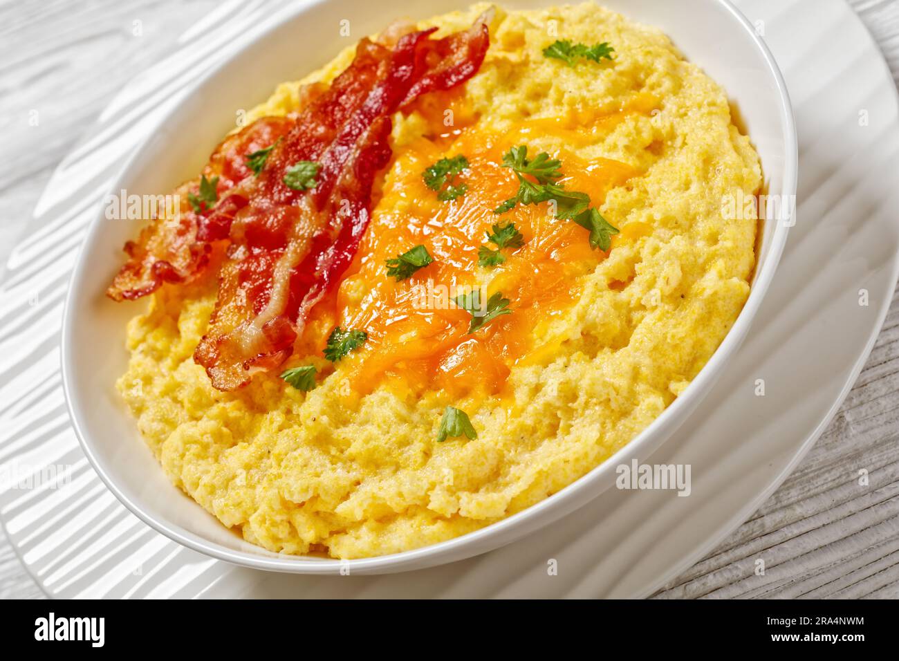 Nahaufnahme von Cheddar Cheese Grits Casserole mit gebratenen Speckscheiben in weißer Schüssel Stockfoto