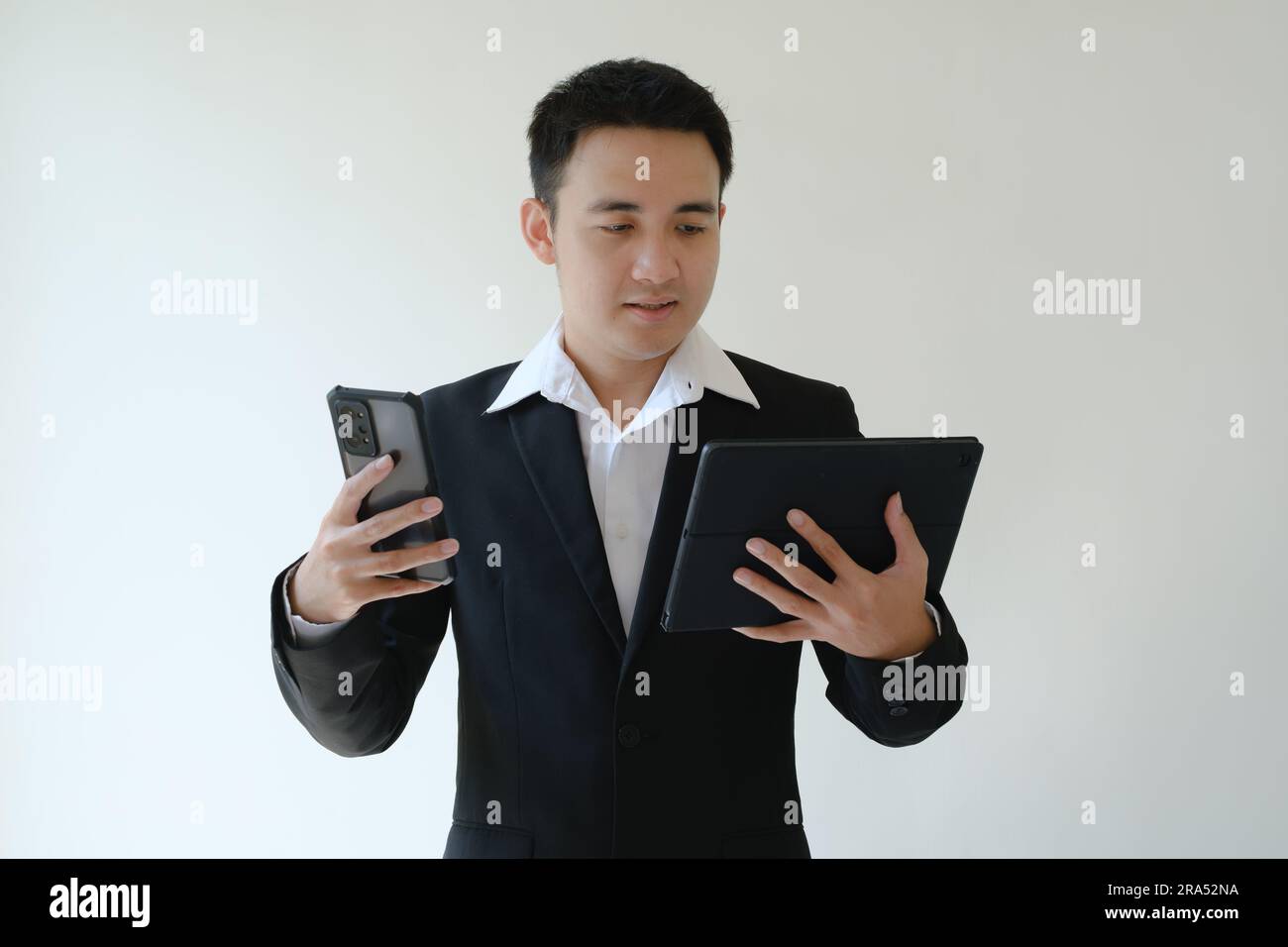 Ein junger asiatischer Geschäftsmann, der ein Smartphone und ein Tablet in der Hand hat und es sich ansieht. Isolierter weißer Hintergrund. Stockfoto