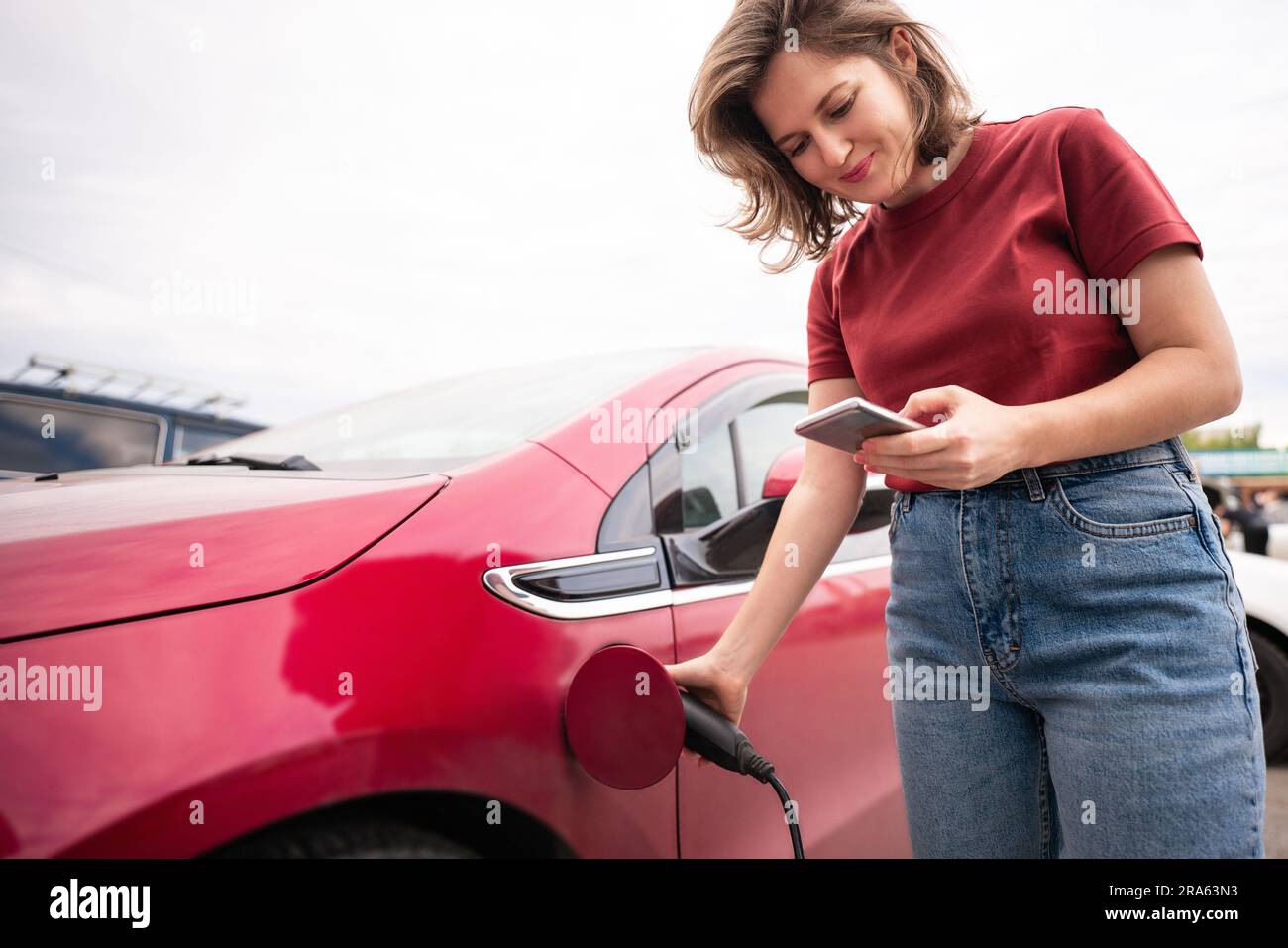 Frau mit Smartphone, die ein rotes Elektroauto auflädt Stockfoto