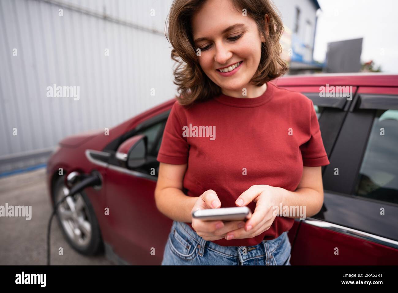 Frau mit Smartphone, die ein rotes Elektroauto auflädt Stockfoto