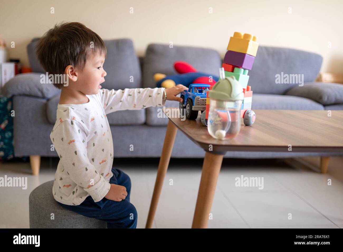 Ein eineinhalb Jahre alter Junge aus gemischten Rassen, der mit bunten Bausteinen und einem formbetonten Spielzeugfahrzeug spielt. Kleinkinder sitzen neben einem Kaffee Stockfoto