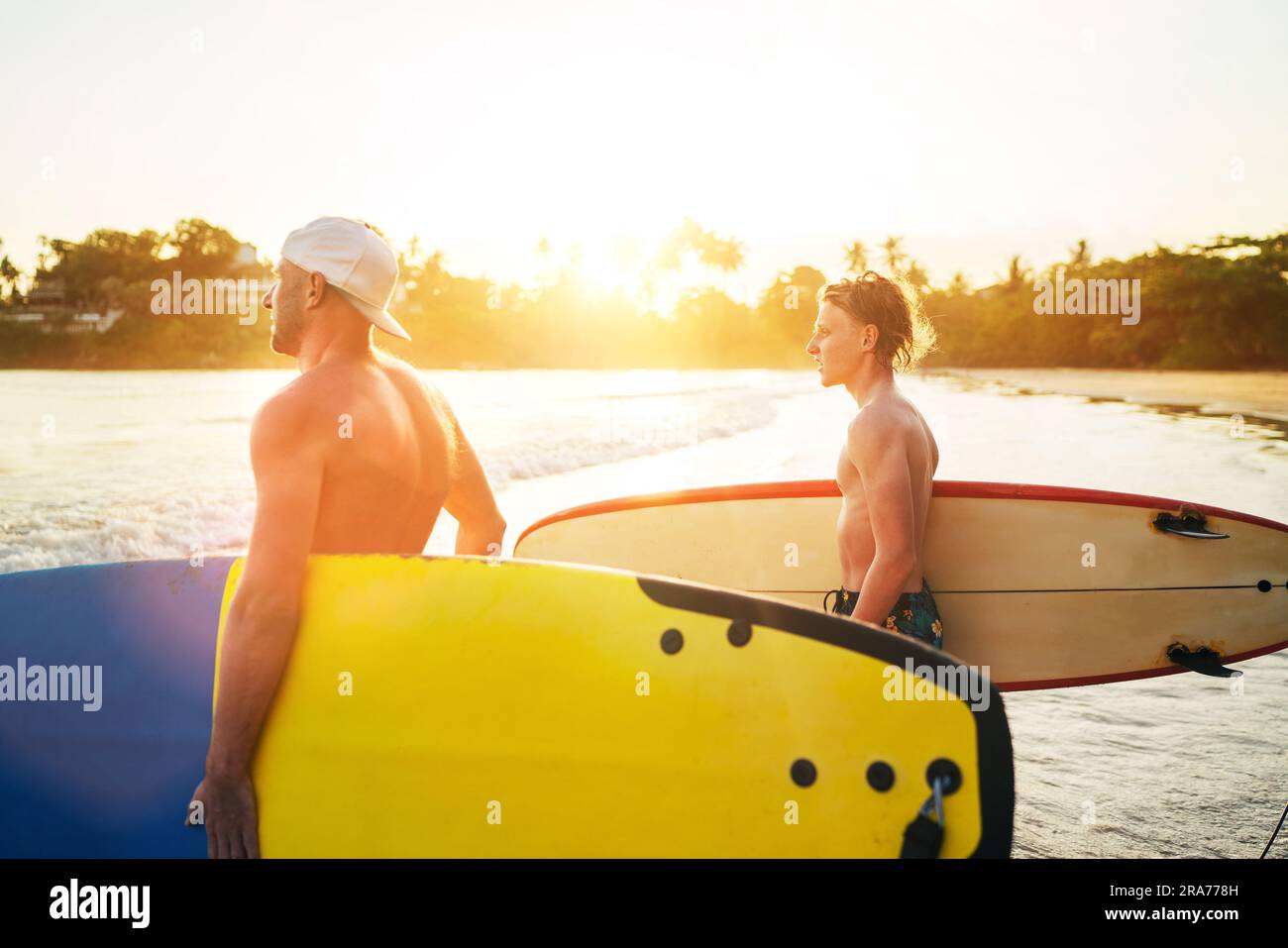 Der junge Teenager mit einem Surfbrett und seinem Vater geht zum Surfen ins Meer. Sie lächeln und genießen ein wunderschönes Sonnenuntergangslicht. Aktiver Familienurlaub Stockfoto
