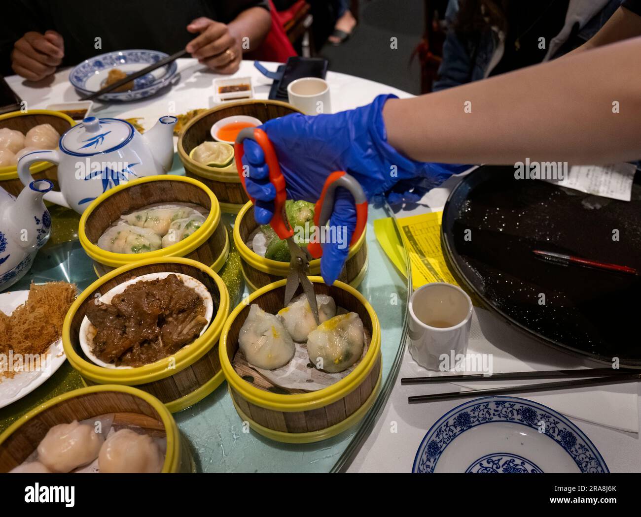 Frauenhand mit Handschuhen, mit Schere und Klößchen auf einem Tisch voller Yumcha-Geschirr. Stift und Bestell Papier auf dem Tisch. Stockfoto