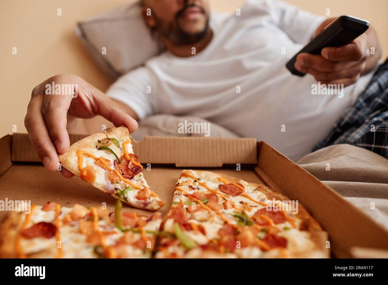 Nahaufnahme eines erwachsenen Mannes, der ein Stück Pizza nimmt, während er ein faules Wochenende zu Hause genießt und Fernsehen und Kopierraum sieht Stockfoto