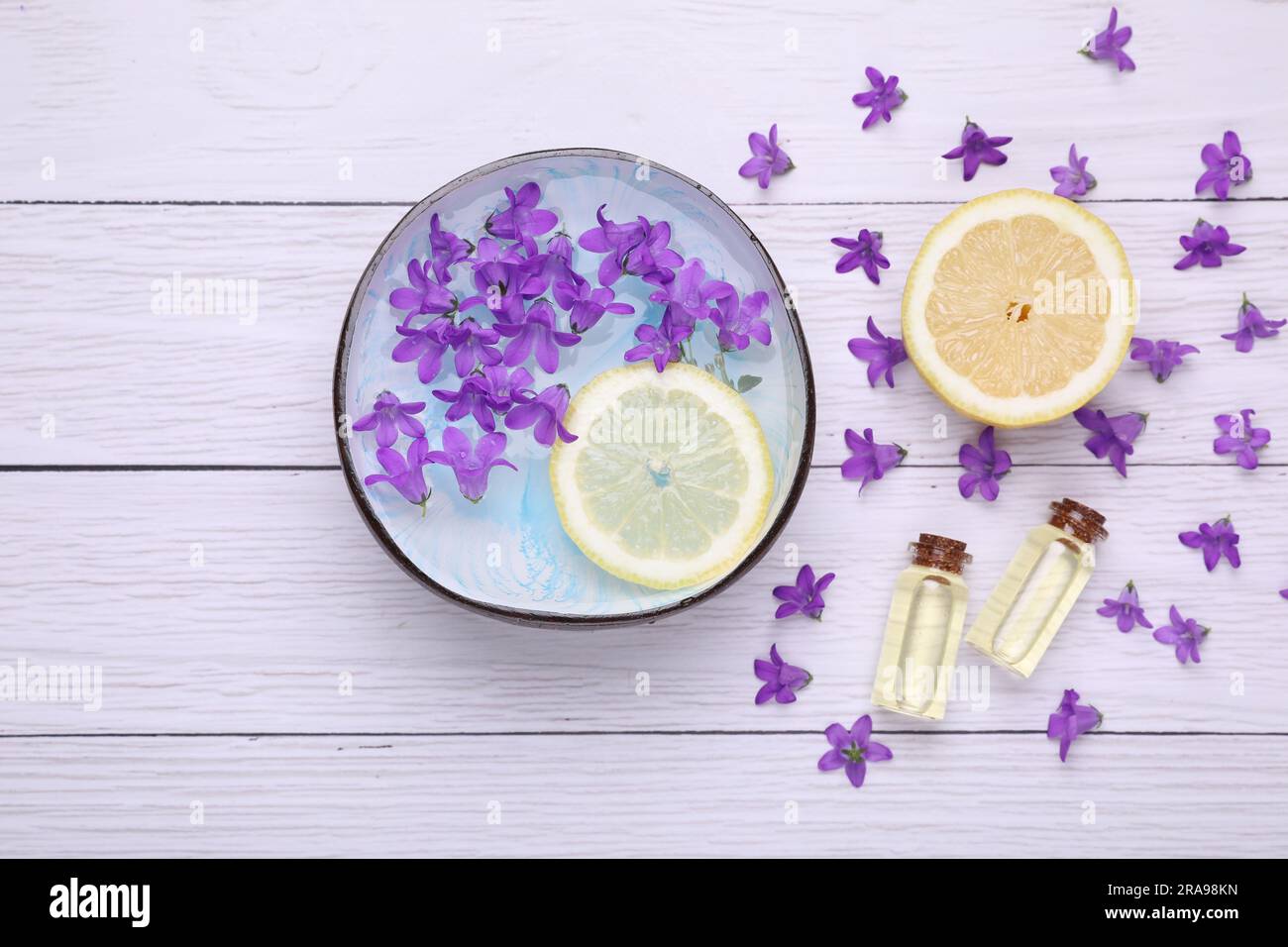 Schüssel mit Wasser, Zitrone, violetten Blumen und Flaschen mit ätherischen Ölen auf weißem Holztisch, flach liegend. Spa-Zusammensetzung Stockfoto