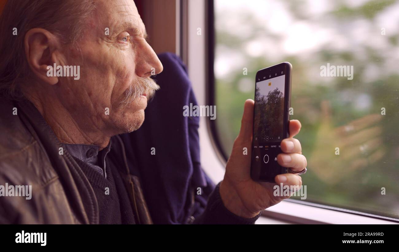Älterer Mann, der mit dem Zug reist und mit einem Smartphone Bilder von der Landschaft durch das Fenster macht Stockfoto