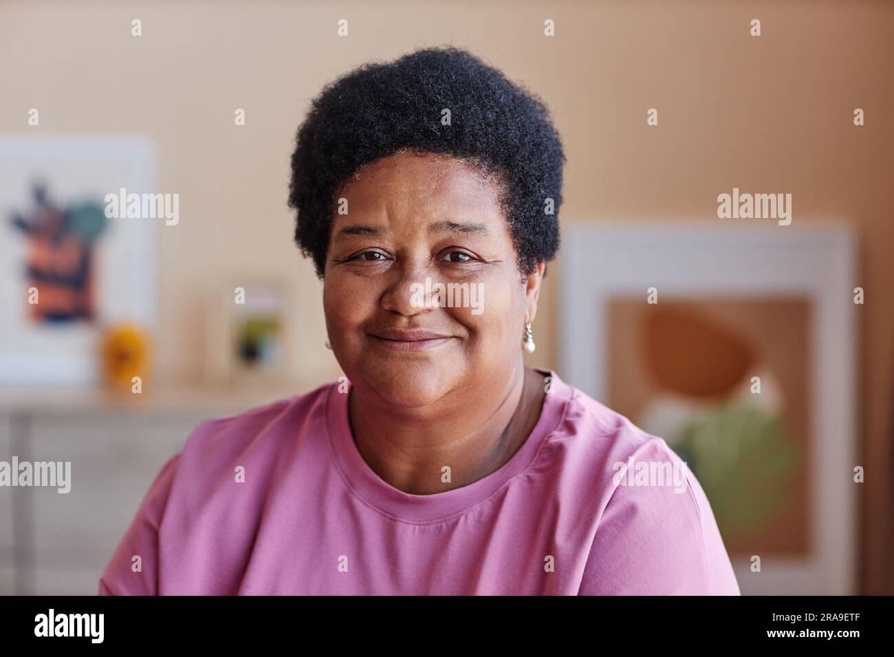 Reife afroamerikanische Großeltern in pinkfarbener Freizeitkleidung schauen in die Kamera, während sie zu Hause gegen Bilder an der Wand sitzen Stockfoto