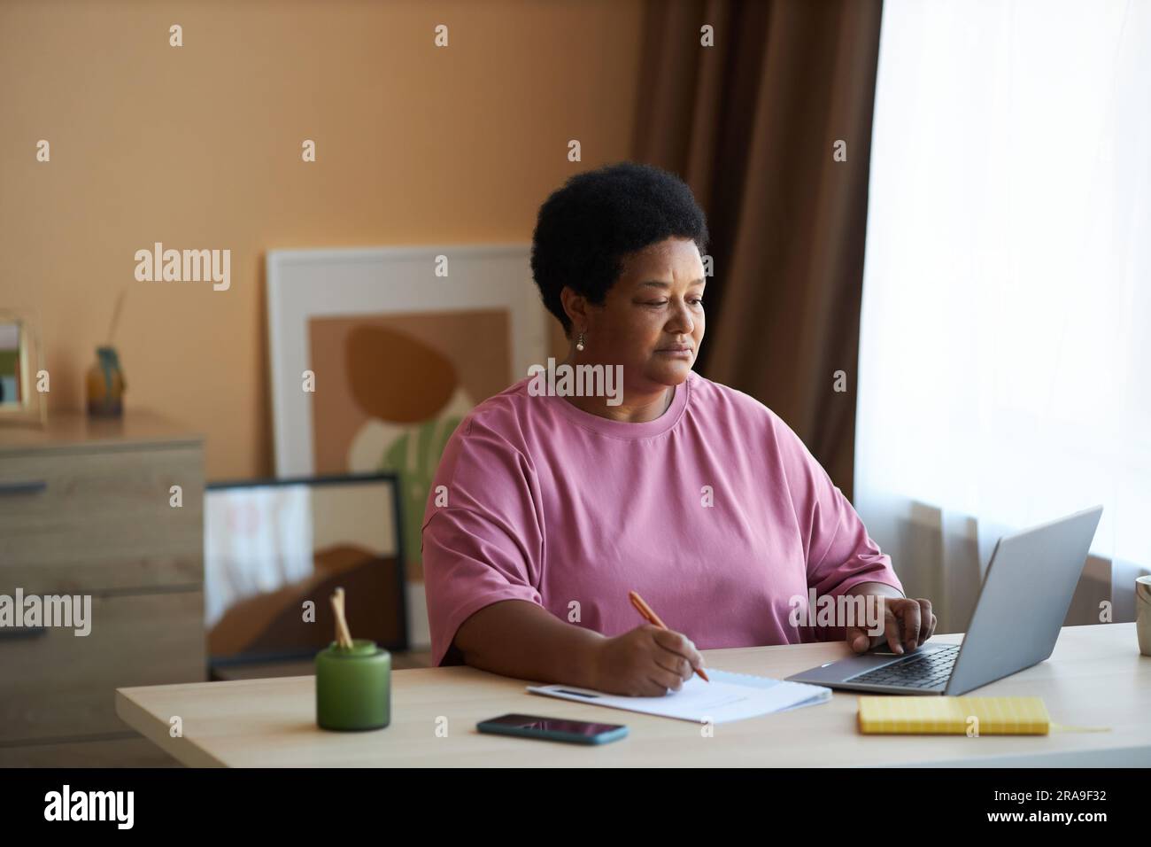Seriöse Geschäftsfrau, die sich Notizen in Dokumenten macht, während sie am Arbeitsplatz vor dem Laptop sitzt und Online-Informationen durchsucht Stockfoto