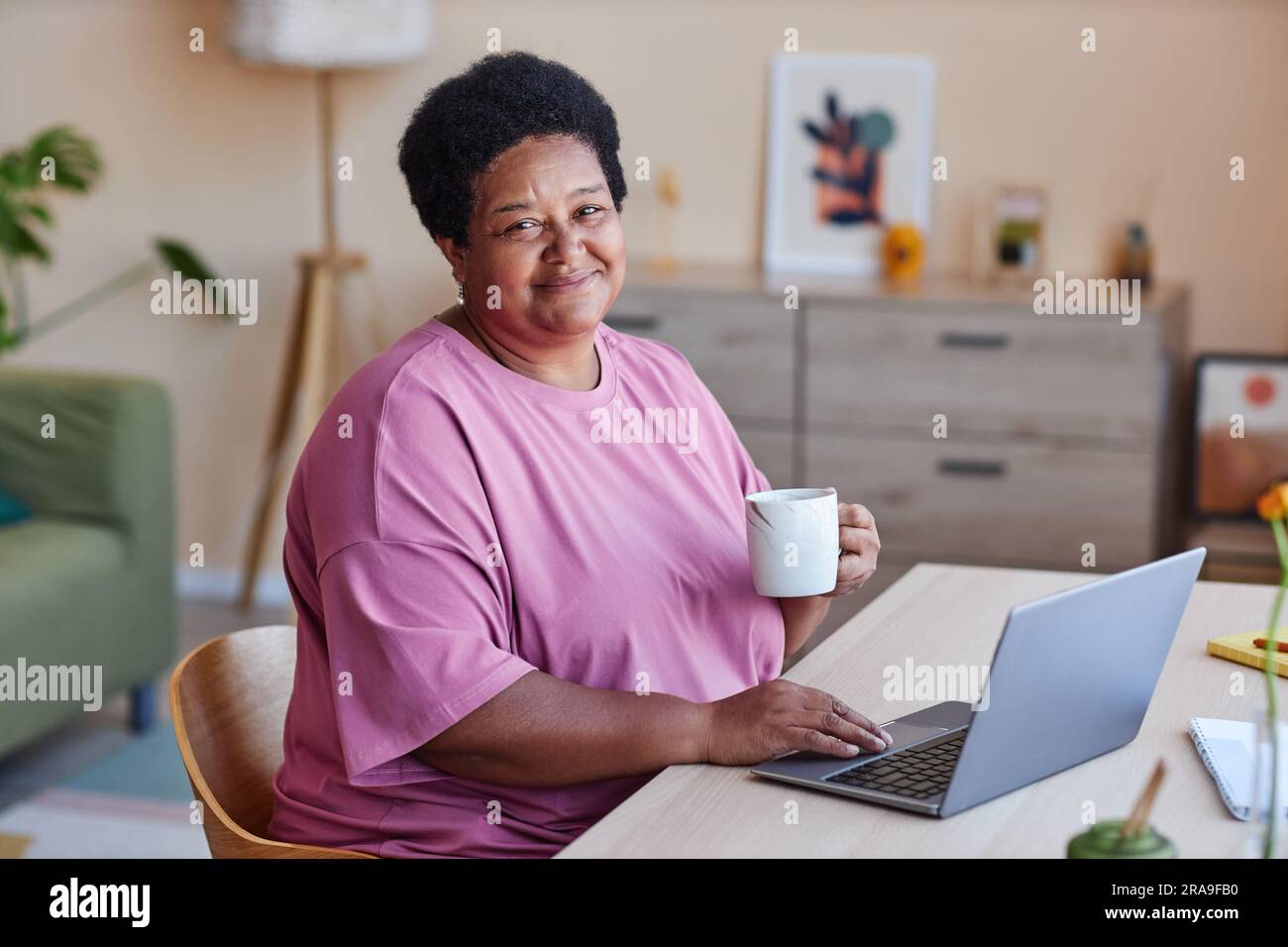 Glückliche Frau mit einer Tasse Tee oder Kaffee, die mit einem Lächeln in die Kamera schaut, während sie vor dem Laptop sitzt und ein Video zum Ansehen auswählt Stockfoto