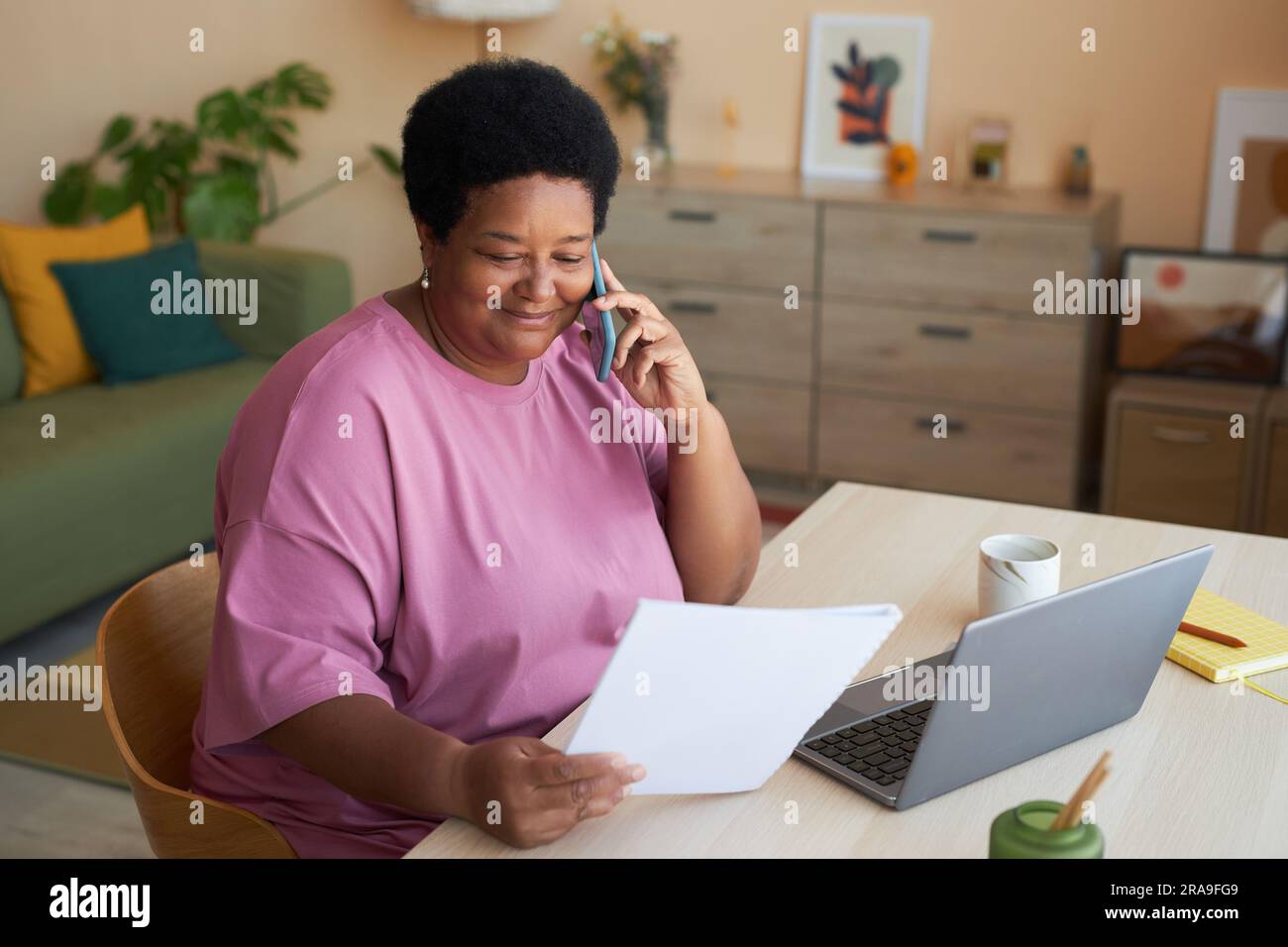 Lächelnde Geschäftsfrau im Alter mit einem Finanzdokument, die mit ihrem Arbeitgeber per Mobiltelefon spricht, während sie am Schreibtisch vor dem Laptop sitzt Stockfoto
