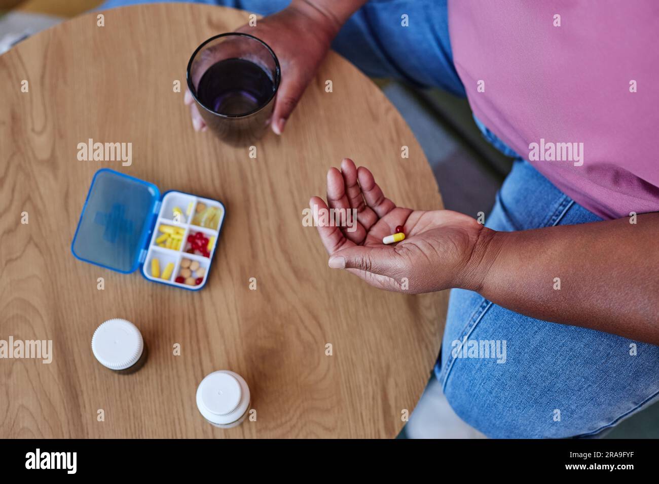 Über dem Winkel einer alten afroamerikanischen Frau in blauen Jeans und rosa T-Shirt, die Pillen nimmt, während sie mit Behälter und Flaschen am Tisch sitzt Stockfoto