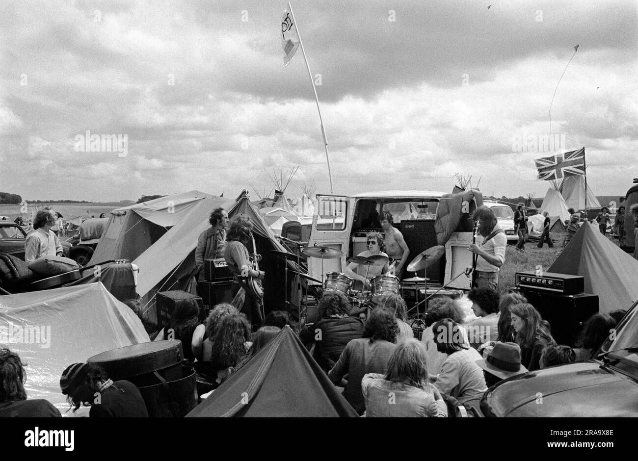 Hippies treten bei einem spontanen Rockkonzert zwischen ihren Zelten auf. Eine kleine Menschenmenge versammelt sich, um zuzuhören. Stonehenge Free Festival bei der Sommersonnenwende, Wiltshire, England, ca. Juni 1976. HOMER SYKES AUS DEN 1970ER JAHREN Stockfoto