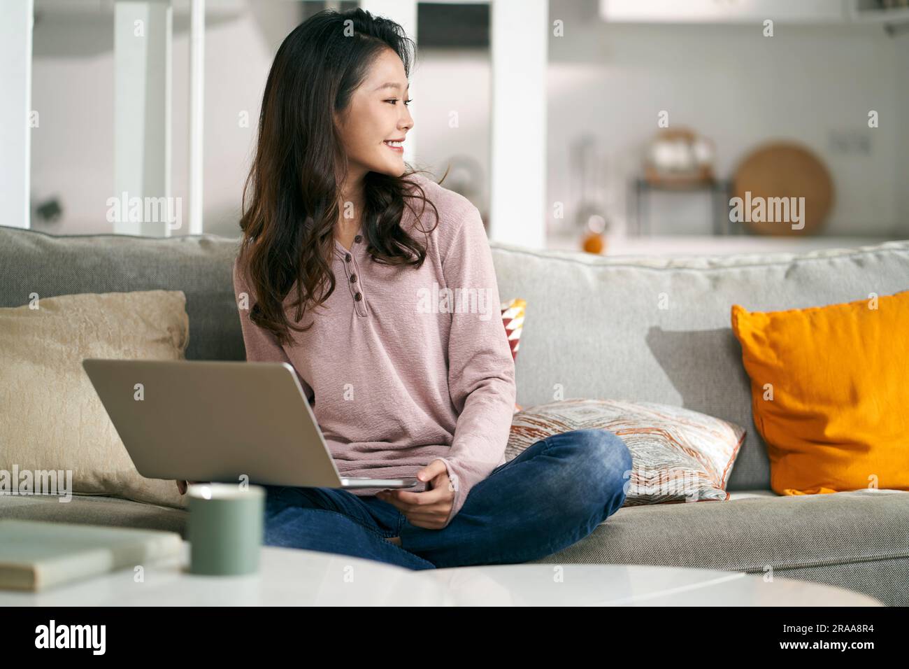 Glückliche junge asiatische Geschäftsfrau, die zu Hause auf der Couch sitzt und mit einem Laptop arbeitet Stockfoto