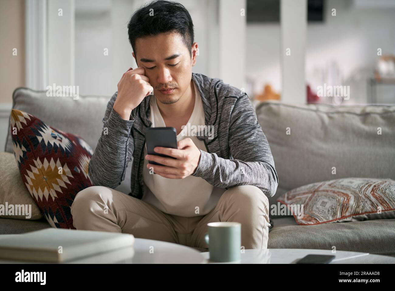 Ein junger asiatischer Mann, der zu Hause auf der Couch sitzt und auf das Handy schaut und ernst aussieht Stockfoto