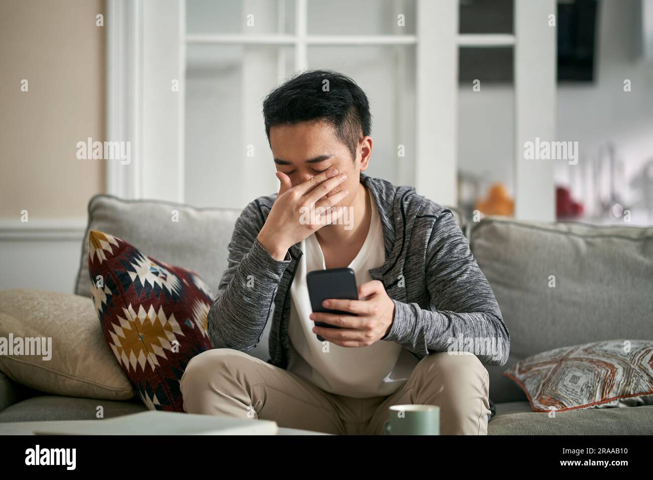 Junger Asiate, der zu Hause auf der Couch sitzt und die Augen mit der Hand verdeckt und sich müde fühlt, nachdem er lange Zeit auf das Handy geschaut hat Stockfoto