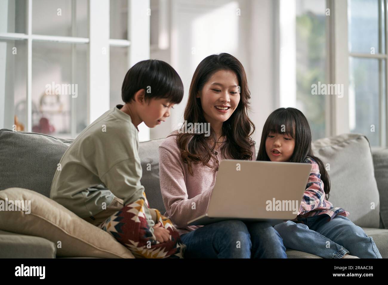 Junge asiatische Mutter und zwei Kinder, die zu Hause auf dem Familientisch sitzen und zusammen einen Laptop benutzen Stockfoto